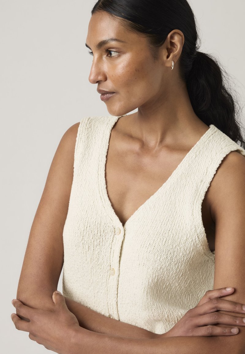 Woman with dark hair in a low ponytail wearing a textured sleeveless cream buttoned top, arms crossed, looking to her left against a plain background.