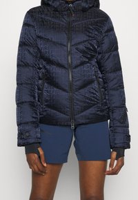 Tmavě modrá péřová bunda s kapucí, zdobená geometrickým vzorem. Má přední zip, boční kapsy a elastické manžety.