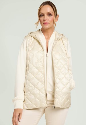 Kobieta ubrana w kremową pikowaną kamizelkę bez rękawów z kapturem, nałożoną na pasującą kurtkę zapinaną na zamek oraz spodnie, stojąca na jasnym, neutralnym tle.