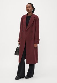 Garšs, divrindas coats bordo krāsā ar ieliektām atloks un melnām pogām. Modele ģērbusies melnā topā un platās biksēs, tur rokās mazu somiņu.