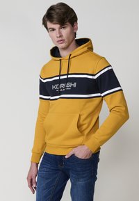 Giovane uomo che indossa un hoodie a righe gialle e navy con tasca a marsupio, in posa su uno sfondo neutro. I jeans blu completano l'outfit.