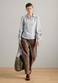 Ανοιχτό μπλε ριγέ πουκάμισο με μακριά μανίκια και φουσκωτά μανίκια, καφέ χαλαρούς παντελόνες, καφέ παπούτσια και γκρί τσάντα ώμου.