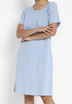 Mujer que lleva un vestido azul claro de manga corta y largo hasta la rodilla con cuello redondo, de pie con los brazos cruzados frente a un fondo blanco.