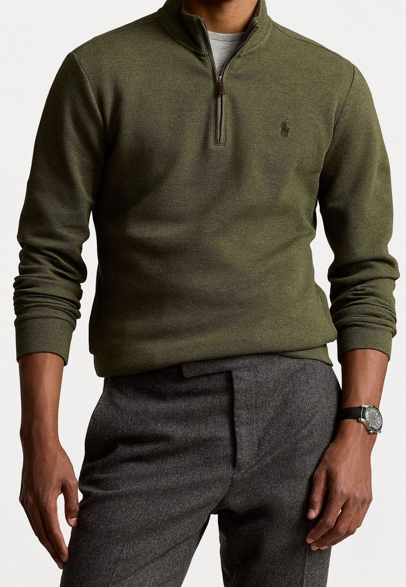 Olivgrüner Pullover mit Reißverschluss am Kragen, gerippten Bündchen und einem dezenten Texturmuster. Getragen mit grauen Hosen und einer Uhr.