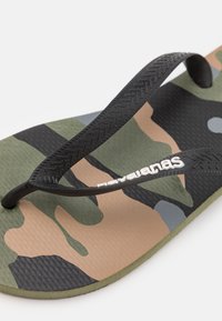 Kamo flip-flop su tekstūruotu juodu guminiu dirželiu. Padas turi žalius, rudus ir pilkus raštus, užtikrinančius sukibimą ir komfortą.