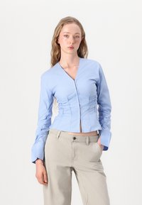 Light blue fitted blouse with a v-neck, gathered detail at the waist, long sleeves, and button closure, paired with beige trousers.