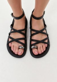 Sandalias negras con tiras cruzadas, hechas de material sintético. Cuentan con una correa para el tobillo y un acento metálico sobre el dedo gordo.