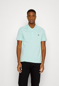 Camisa polo de color azul claro con cuello, mangas cortas y un pequeño logo bordado en verde. Llevada con pantalones negros, exhibida sobre un fondo neutro.