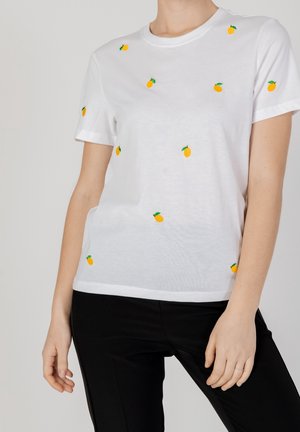 Camiseta blanca de algodón con pequeños limones amarillos bordados y hojas verdes, cuello redondo y mangas cortas. Combinada con pantalones negros.