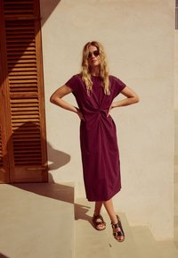 Robe violette longueur genoux avec un design noué à l'avant. Confectionnée en tissu doux, manches courtes, associée à des sandales marron avec des détails dorés.