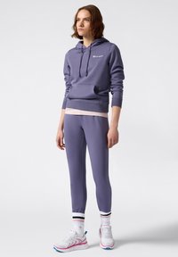 Sudadera morada clara con un bolsillo frontal, combinada con leggins a juego. Presenta una capucha, puños acanalados y un logo blanco. Zapatillas blancas con acentos rosas.