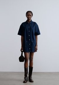 Tengerészkék, gombos lenge ruha rövid ujjal, két elülső zsebbel, fekete térdig érő csizmával és egy kisméretű fekete táskával kombinálva. Sima, texturált anyag.