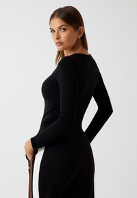 Woman with straight brown hair wearing a fitted black long-sleeve top and black pants with white and brown side stripes, hands in pockets, looking back.