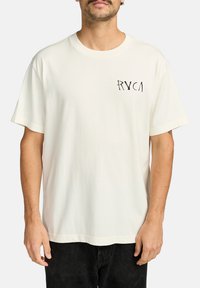 Camiseta de manga corta color crema, hecha de algodón, con un ajuste relajado, que presenta un pequeño logo gráfico negro en la zona del pecho izquierdo.