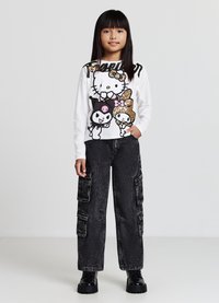 Camicia bianca a maniche lunghe con personaggi dei cartoni animati colorati e testo; abbinata a pantaloni cargo neri con molte tasche e tessuto testurizzato.