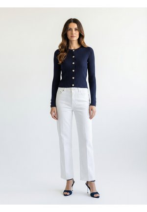 Frau steht und trägt eine marineblaue, gerippte, geknöpfte Strickjacke, weiße, hoch taillierte, gerade geschnittene Hose und marineblaue Sandalen mit hohen Absätzen vor weißem Hintergrund.