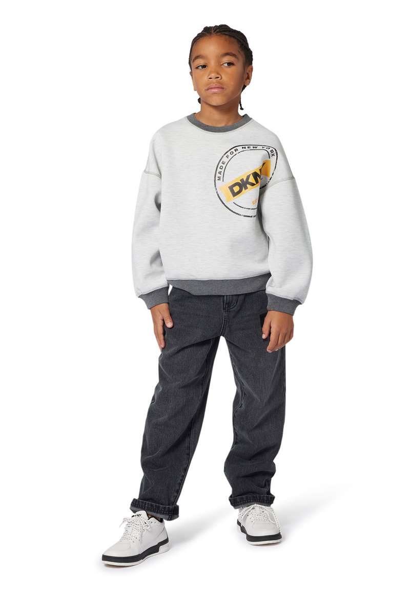 Sweatshirt gris avec un logo rond et des accents jaunes, associé à un jean foncé et des baskets blanches. Caractéristique d'une coupe décontractée et de manches retroussées.