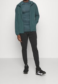 Chaqueta ligera color verde azulado con cremallera, camiseta ceñida verde azulado, pantalones deportivos negros y zapatillas deportivas negras. Incluye capucha ajustable y bolsillos laterales.