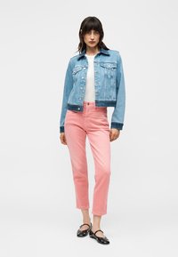 Femeie în picioare, purtând o geacă din denim albastru deschis, top alb, pantaloni roz scurți și pantofi Mary Jane negri pe un fundal alb simplu.