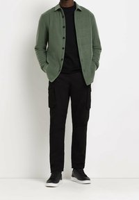 Zielona polarowa kurtka na guziki noszona na czarną koszulę, w zestawieniu z czarnymi spodniami cargo i czarnymi trampkami z białymi podeszwami.