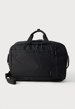 Sac de voyage rectangulaire noir avec doubles poignées, poche avant zippée et bandoulière réglable sur fond gris clair.