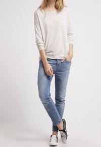 Ljusbeige långärmad topp med avslappnad passform, tillsammans med medelblå skinny-jeans. Silver- och svartmönstrade slip-on-skor.