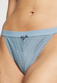 Lichtblauwe kanten string met een zachte tailleband, voorzien van een kleine strik en decoratieve accenten, die een textuurpatroon en ronde randen laat zien.
