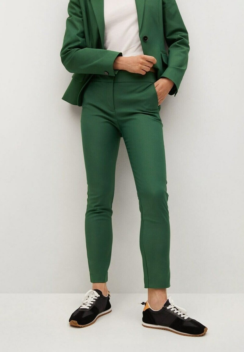 Personne portant un pantalon de costume vert ajusté et un blazer avec une chemise blanche et des baskets noires à lacets blancs, debout contre un mur blanc.