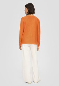 Oranges Strickpullover mit entspannter Passform, rundem Ausschnitt und langen Ärmeln, kombiniert mit weit geschnittenen, hellcremefarbigen Hosen und beigen Schuhen.