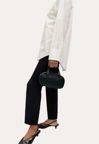 Persoon in een witte lange mouw shirt en zwarte broek, houdt een kleine zwarte handtas vast, draagt zwarte lage slingback schoenen.
