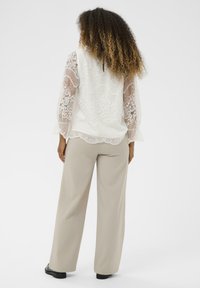 Blouse en dentelle avec des manches longues transparentes, motifs floraux blancs et ourlet festonné. Associée à un pantalon large beige et des chaussures noires.