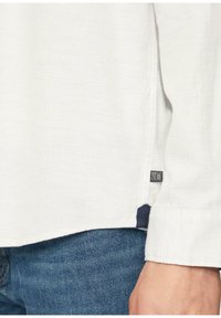 Witte, textuurachtige lange mouwen shirt met manchet en zoom met klein label, gedragen met blauwe spijkerbroek en een rustende hand zichtbaar aan de zijkant.