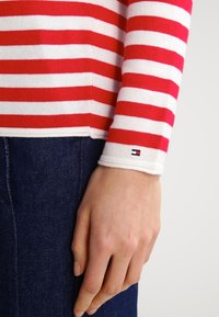 Rød- og hvidstribet langærmet bluse med ribbet kant, der har et lille logo på manchetterne, parret med mørk denim.