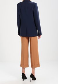 Tmavě modrý saka na míru s vycpanými rameny, doplněné o vysoké kalhoty se širokými nohavicemi v barvě světle hnědé, s ohrnutými manžetami a černými podpatky.