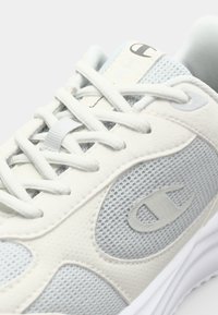 Baskets grises claires avec une tige en mesh, une languette rembourrée et des accents texturés. Comprend des lacets plats et un logo en relief sur le côté.