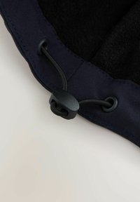 Reguliuojama juoda elastinga virvė su plastikiniu užtrauktuku, priveržta per dvi akutes tamsaus audinio krašte, prieš šviesų foną.