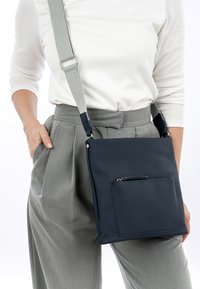 Borsa a tracolla blu navy realizzata in materiale liscio. Presenta una tasca frontale con zip, una tracolla grigia regolabile e un design minimalista.