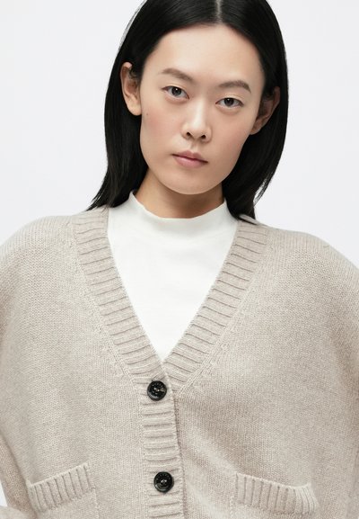 Cardigã bege de malha com decote em V, bordas caneladas, três botões pretos e dois bolsos frontais, usado sobre um gola alta branco.