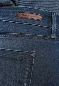 Tmavé denimu kalhoty s hnědou koženou etiketou s nápisem "Maui Jeans Co." na pase a kovovým nýtem s písmenem "M" na zadní kapse.