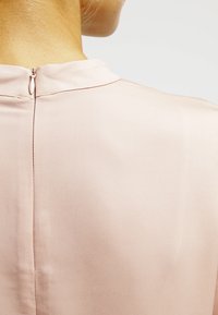 Jasnoróżowa bluzka o miękkiej, gładkiej fakturze; ma wysoki dekolt i krótki zamek z tyłu, co podkreśla prostotę w designie.