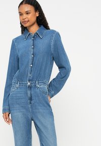 Denim jumpsuit in light blue with a button-up front, collar, long sleeves, and cinched waist. Features pockets and subtle fading.