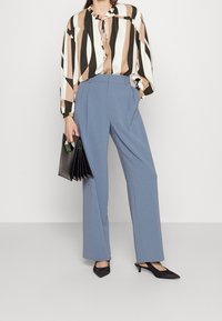 Modré široké kalhoty se záhyby, doplněné vzorovanou blůzou v krémové, černé a hnědé barvě. Outfit doplňuje černé skládací psaníčko.