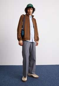 Veste zippée marron sur une chemise rayée bleu clair, pantalon large gris et sneakers beiges avec accents marron. Complété par une casquette verte.