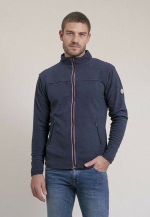 Homme portant une veste polaire bleu marine zippée avec poches latérales et un jean bleu, debout devant un fond clair uni.