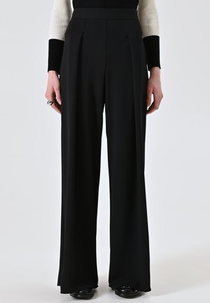 Pantaloni neri a gamba larga in un tessuto liscio con pieghe frontali, dotati di una vita alta e una vestibilità sartoriale. Le scarpe nere completano il look.