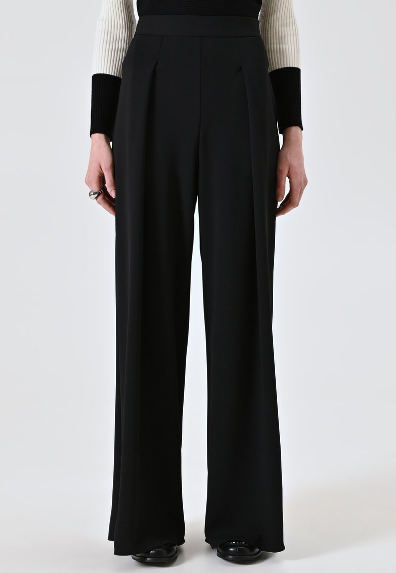 Pantaloni neri a gamba larga in un tessuto liscio con pieghe frontali, dotati di una vita alta e una vestibilità sartoriale. Le scarpe nere completano il look.