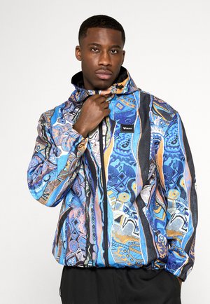 Hombre con una chaqueta con cremallera y capucha de estampado azul, naranja y negro, sujetando la chaqueta cerca de su barbilla.