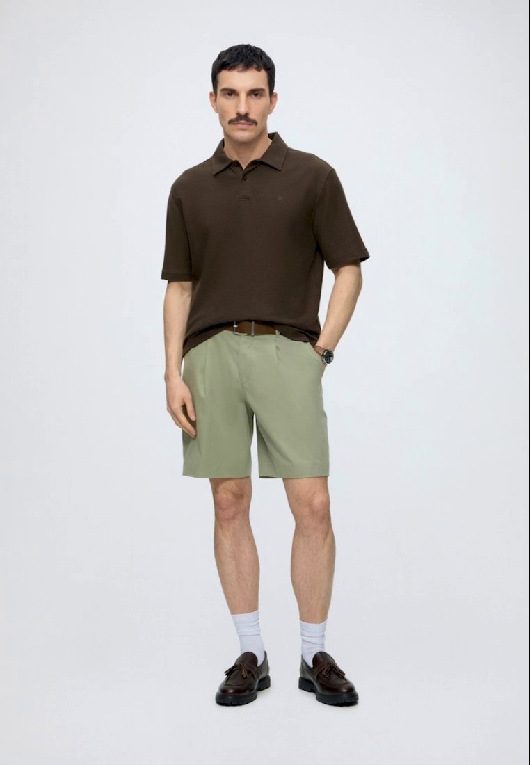 Homme debout portant un polo marron foncé, un short vert clair avec ceinture, des chaussettes blanches et des chaussures marron foncé sur fond uni.