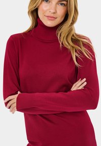Ribbed turtleneck sweateri syvän punaisena, jossa on pitkät hiat ja istuva muotoilu. Pehmeä rakenne, jossa on virtaviivainen muoto ja minimaalinen yksityiskohtaisuus.