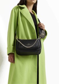 Borsa a tracolla in pelle nera con catena, caratterizzata da una superficie liscia e un logo impresso. Indossata insieme a un cappotto verde brillante.